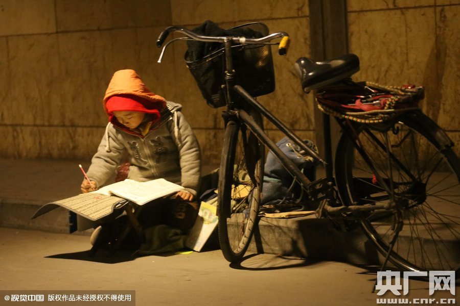 鄭州中原路京廣鐵路大橋下涵洞的馬路牙子邊，一位戴著紅色辣椒帽的小男孩趴在一個(gè)小馬扎上，上面鋪著一張車墊，在微弱的燈光下寫作業(yè)